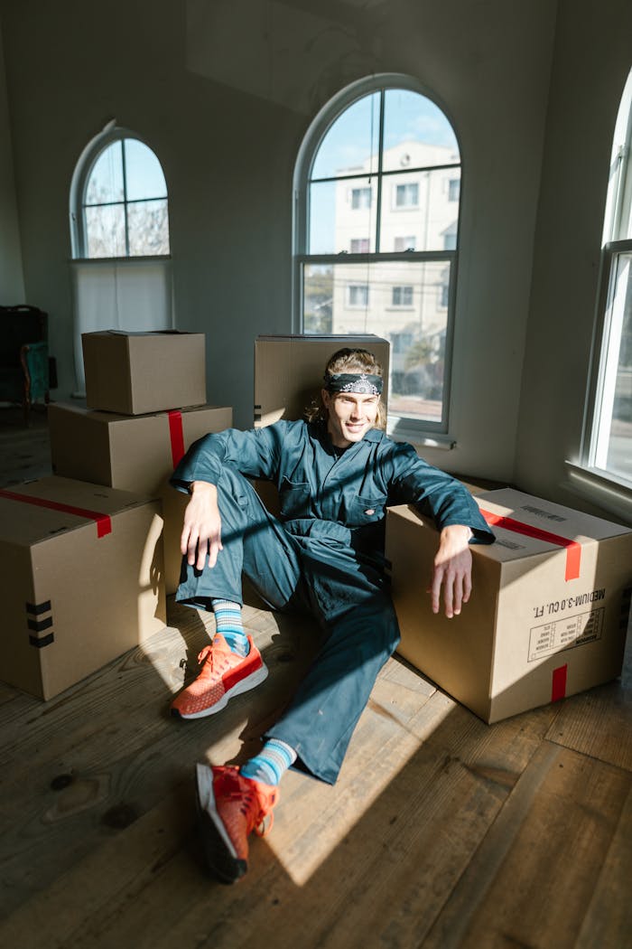 Man sitting among moving boxes in a sunlit room, wearing coveralls and looking relaxed.